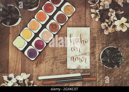 Watercolour paints on a table with pot plants, flowers and a card with the words hecho con amor Stock Photo