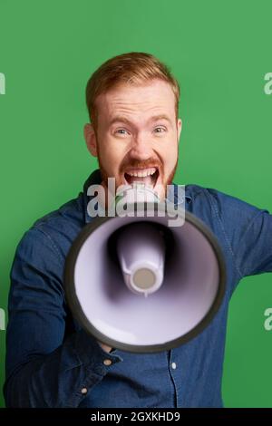 Portrait of emotional young Caucasian man with red beard making announcement through loudspeaker Stock Photo
