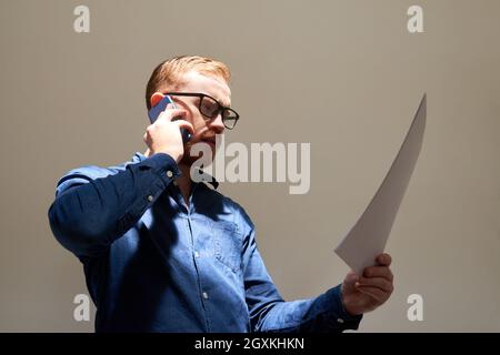 Below view of frowning young Caucasian businessman in glasses talking by phone and analyzing papers Stock Photo
