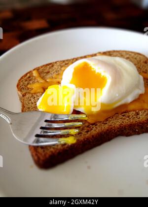 Poached egg with yolk broken onto toast on a white plate against a dark background Stock Photo