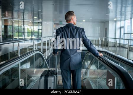 Man with suitcase on escalator with back to camera Stock Photo