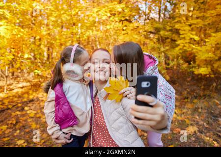 a cheerful woman takes selfies with her daughters in an autumn park