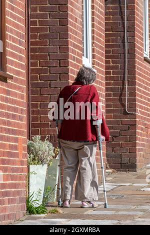 elderly lady on crutches struggling to walk up a hill next to some red brick houses. disabled old lady with walking crutches on hill. Stock Photo