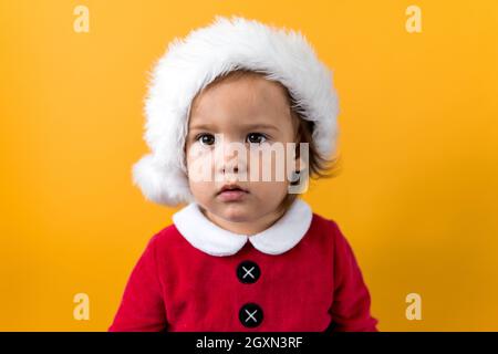 Serious Portraite Cute Happy Cheerful Chubby Baby Girl in Santa Hat Looking On Camera At Yellow Background. Child Play Christmas Scene Celebrating Stock Photo