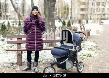 Portrait of young adult caucasian happy mom in beautiful purple warm down jacket using smartphone device walking with newborn baby stroller at city Stock Photo