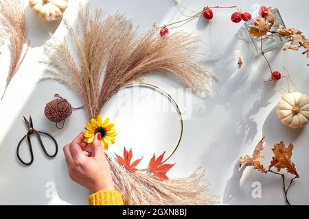 Hands making dried floral wreath from dry pampas grass and Autumn leaves. Hands in sweater with manicured nails tie decorations to metal frame. Flat Stock Photo