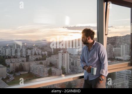 young man enjoying evening coffee and beautiful sunset landscape of the city while standing by the window Stock Photo