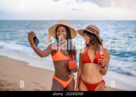 Happy slim young multiracial female best friends in bikinis and hats with glasses of cocktail having fun and taking selfie on smartphone during summer holidays on seashore Stock Photo