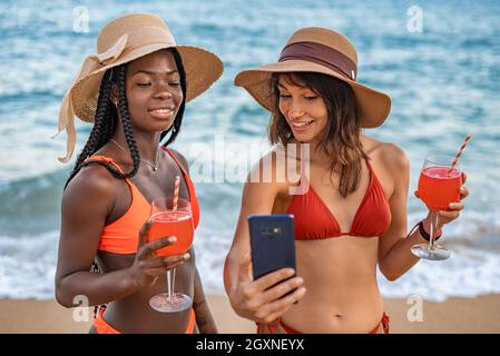 Stylish happy young diverse female friends in bikinis and hats holding glasses with red drinks and taking selfie on mobile phone while chilling together on seashore in summer day Stock Photo