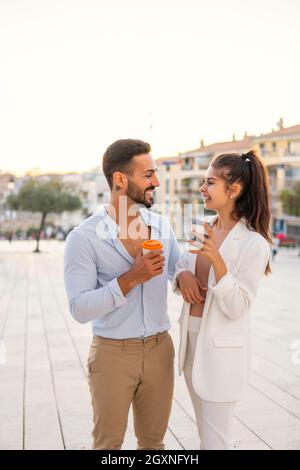 Positive stylish Hispanic couple with cups of takeaway coffee standing on square in city and looking at each other during summer stroll in sunset Stock Photo