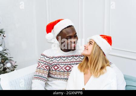 Happy New Year and Merry Xmas at home together. Smiling young african american guy and caucasian lady in Santa Claus hats, hug and look at camera in l Stock Photo