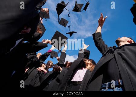 Congratulations! Low angle view of happy group of six young cheerful graduates in black gowns, throwing up their head wear in the air and celebrating, Stock Photo