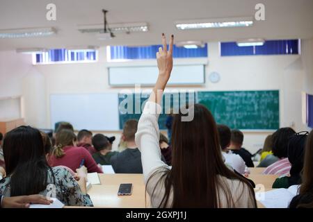 Rear view of female student sitting in the class and raising hand up to ask question during lecture. High school student raises hand and asks lecturer Stock Photo