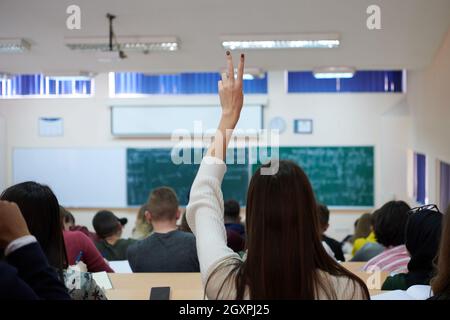 Rear view of female student sitting in the class and raising hand up to ask question during lecture. High school student raises hand and asks lecturer Stock Photo