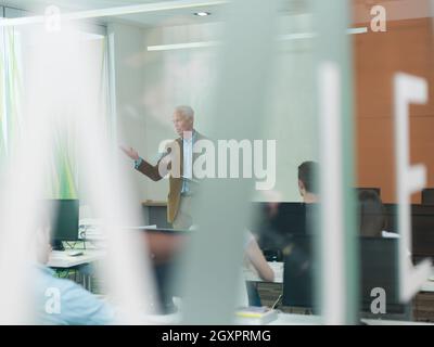 Handsome senior teacher and students group in computer lab classroom. Experienced professor using tablet computer while presenting lessons to class. Stock Photo