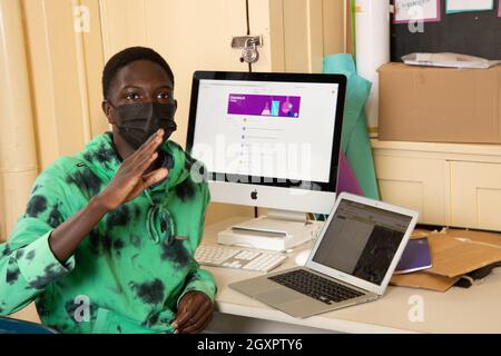 Education High School classroom scene, male student talking in class, gesturing, with desktop and laptop computer on desk, wearing face mask Stock Photo