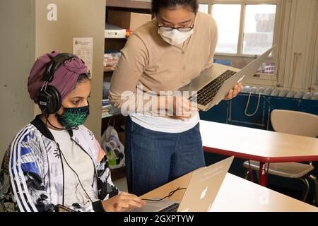 Education High School classroom scene female teacher leaning over to assist female student using laptop computer in class, both wearing face masks Stock Photo