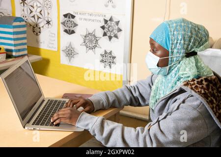 Education High School classroom scene female student in head scarf working at laptop computer in classroom, wearing face mask to protect vs Covid-19 Stock Photo