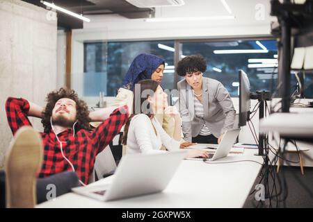 young software developer resting with legs on desk while his multiethnics business team writing programming code in the background at startup office Stock Photo