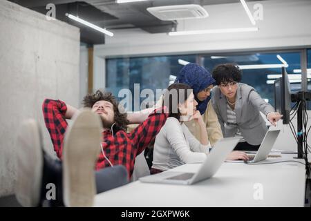 young software developer resting with legs on desk while his multiethnics business team writing programming code in the background at startup office Stock Photo