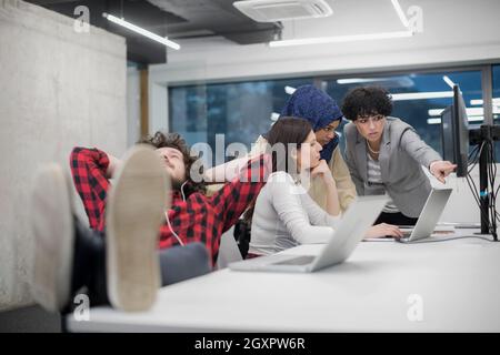 young software developer resting with legs on desk while his multiethnics business team writing programming code in the background at startup office Stock Photo