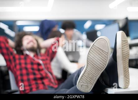 young software developer resting with legs on desk while his multiethnics business team writing programming code in the background at startup office Stock Photo