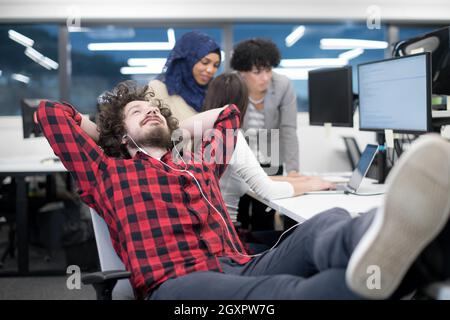 young software developer resting with legs on desk while his multiethnics business team writing programming code in the background at startup office Stock Photo