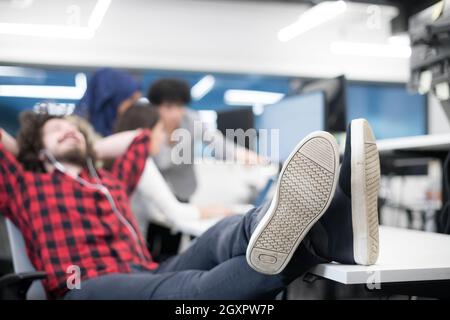 young software developer resting with legs on desk while his multiethnics business team writing programming code in the background at startup office Stock Photo