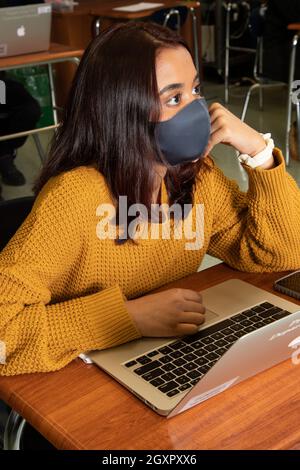 Education High School, classroom scene, female student listening in class, laptop computer on desk in front of her, wearing face mask Covid-19 Stock Photo