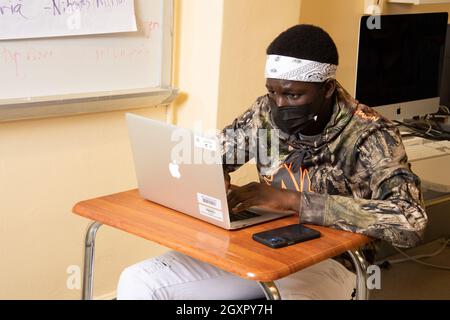 Education High School male student at work on laptop computer in classroom, computer monitor behind him, wearing face mask to protect against Covid-19 Stock Photo