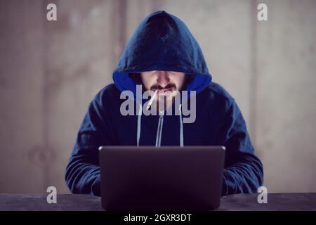Young talented hacker smoking cigarette while working on laptop computer in dark office with concrete wall in the background Stock Photo