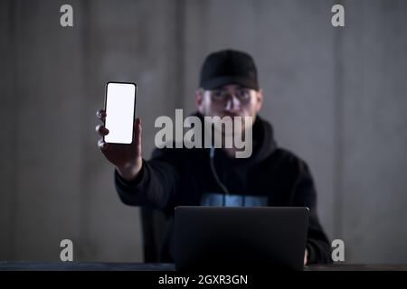 Young talented criminal hacker using laptop computer and showing the smart phone which is pirated while working in dark office with concrete wall in t Stock Photo