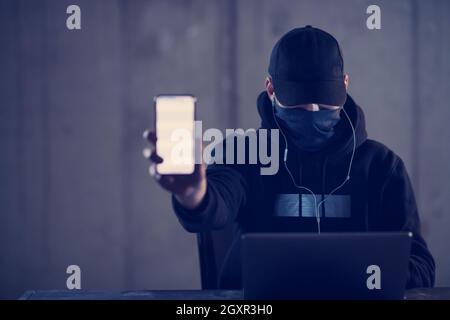 Young talented criminal hacker using laptop computer and showing the smart phone which is pirated while working in dark office with concrete wall in t Stock Photo