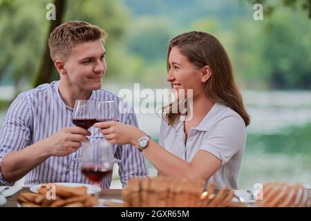 young happy couple toasting red wine glass while having picnic french dinner party outdoor during summer holiday vacation near the river at beautiful Stock Photo