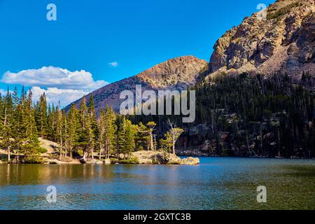 Pine trees next to large lake in the mountains with blue sky Stock Photo