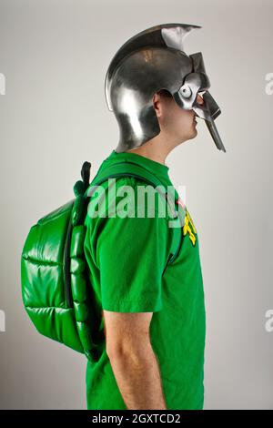 Profile of a white man wearing a green shirt, turtle shell backpack, and Trojan battle helmet Stock Photo