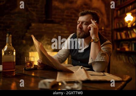 Man smokes cigarette, drinks alcohol beverage and reads newspaper, bookshelf and vintage office interior on background. Tobacco smoking culture, speci Stock Photo