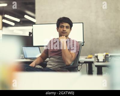 Portrait of young indian successful male software developer sitting on desk at modern startup office Stock Photo