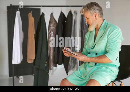 Stylish middle aged man wearing colorful outfit using tablet pc while sitting next to clothes rail in studio Stock Photo