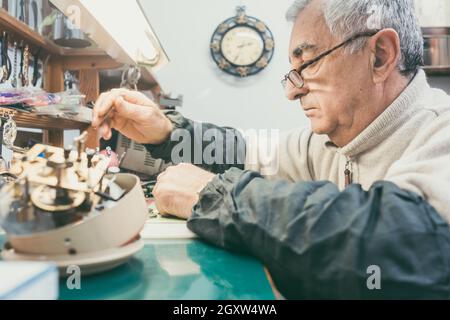 Watchmaker in his workshop repairing a larger clock with clockwork in foreground Stock Photo