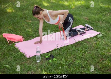Young woman with ponytail in tracksuit does exercises with elastic band kneeling on field Stock Photo