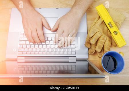 Work Gloves, Level and Coffee Cup Next To Contractor Reviewing Project on Laptop. Stock Photo