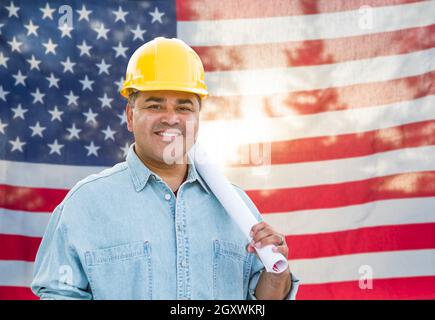 Hispanic Male Contractor with Blueprint Plans Wearing Hard Hat In Front of American Flag. Stock Photo