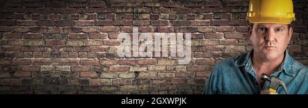 Male Contractor In Hard Hat Holding Hammer In Front Of Old Brick Wall Banner with Copy Space. Stock Photo