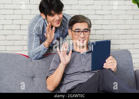 Asian senior businessman with digital tablet discuss together with young team in office. Father man and his son sit on sofa talking chatting on video Stock Photo