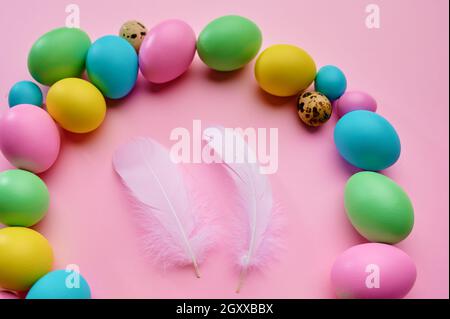 Colorful easter eggs and two white bird feathers on pink background, top view. Paschal food, event decoration, spring holiday celebration symbol Stock Photo