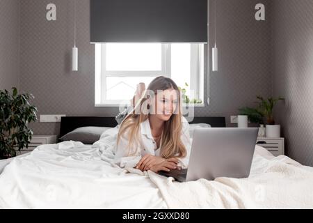 Pretty smiling Caucasian girl lays on bed in white-gray bedroom and use laptop for remote work, talking on video call. Communication, technology, remo Stock Photo