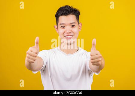 Asian handsome young man smiling positive showing thumbs up gesture good or like sign looking to camera, studio shot isolated on yellow background Stock Photo