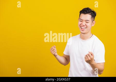 Asian handsome young man smiling positive shaking hands enthusiastic shouting yes for win competition, male raising his fists with smiling delighted f Stock Photo