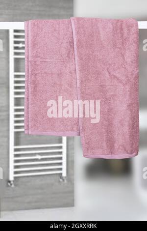 Hanging towels. Closeup of violet pink soft terry bath towels hang on a clothes rail in front of abstract blurred bright gray bath background. Stock Photo